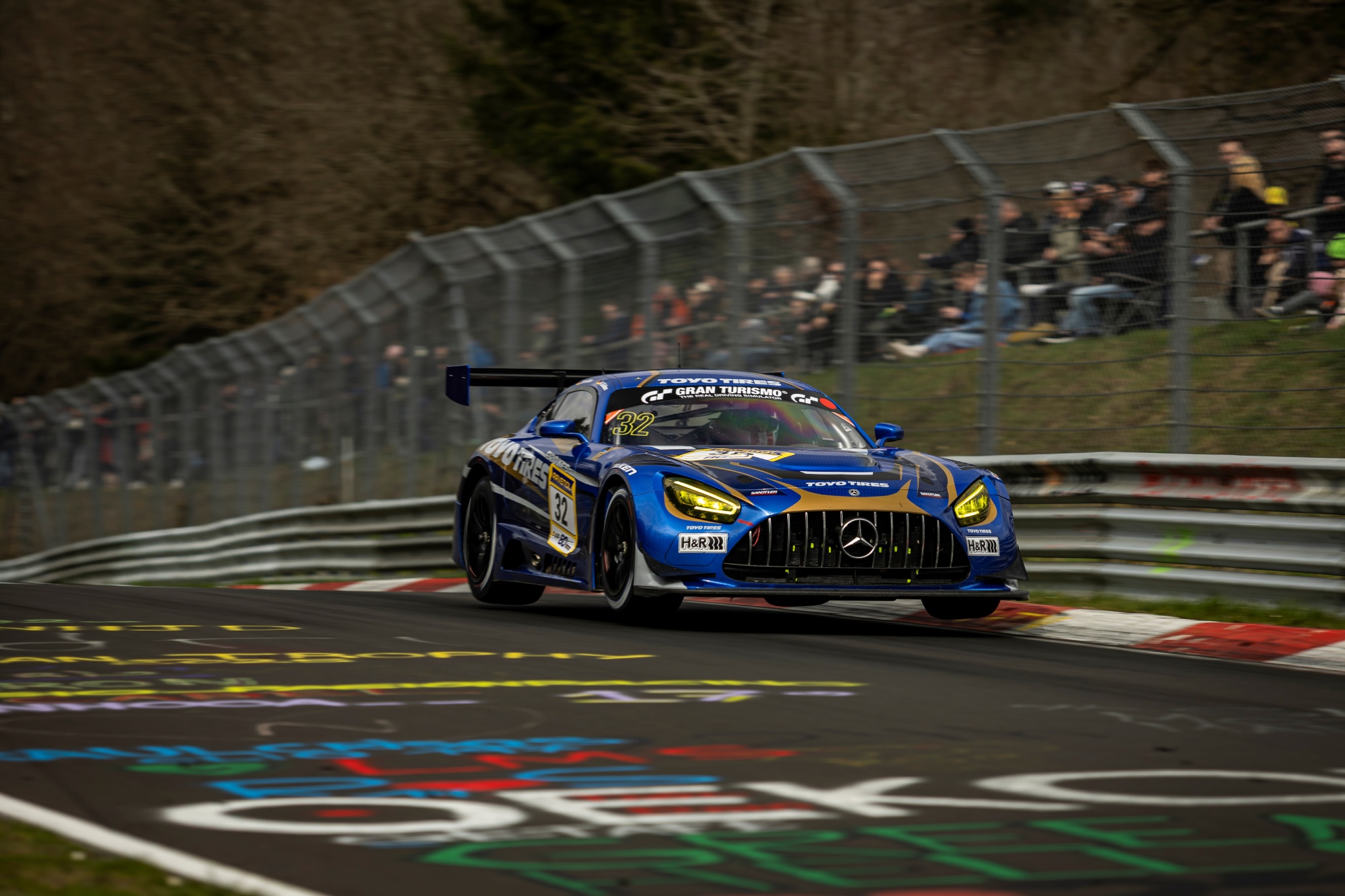 Toyo Tires branded race car is driving on the Nürburging race track and many spectators are behind the fence celebrating the 24h race cars